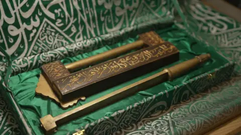 A brass coloured lock and key inscribed with Arabic writing. It is placed in a rectangular box which is covered in green and white cloth.
