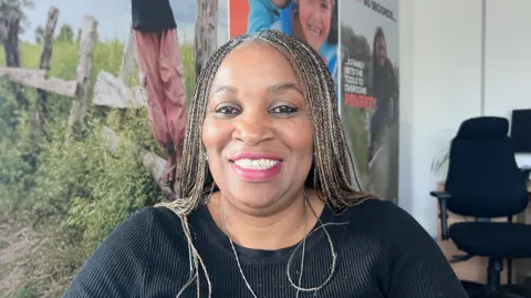 Fola Komolafe is seated indoors in an office setting. She has long braided hair and is wearing a long‑sleeved, ribbed black top. Behind her is a large printed backdrop featuring outdoor scenes with wooden fencing, greenery, and people standing in nature. To one side of the image, there is a standard office chair and a desk with a small plant on it. 