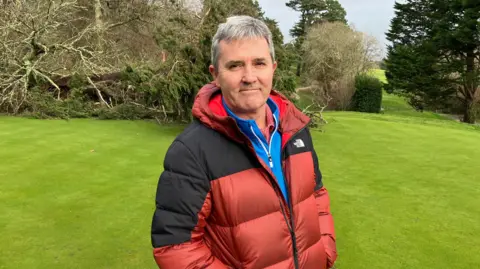 A man wearing a red and black puffer coat, looking towards the camera. He is stood on grass with a large fallen tree behind him.
