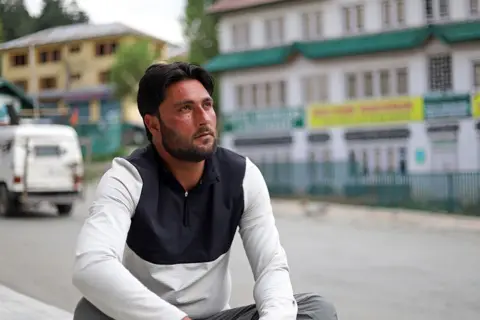 SYED SHAHRIYAR / BBC A man sitting on the side of a street with buildings in the background.
