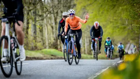 Paul Campbell A cyclist wearing an orange top waves to the camera as they cycle up a tree-lined rural road.