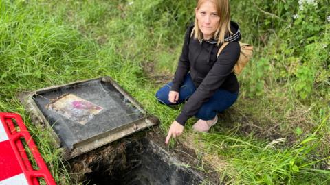 Georgina Heyburn with long blond hair wearing black top and pointing at an open manhole