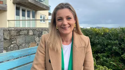 BBC Ellie Lawrence is sitting outside on a blue wooden garden bench and she is smiling at the camera. She has long blonde hair and is wearing a white top, a bright green jacket, a pale brown coat and large gold and pearl earrings. Behind her is a building with French windows and a balcony and to her left are tall shrubs. In the far distance a part of a headland can be seen.