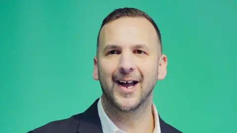 PA Media Zack Polanski Green party leader. a man with his mouth open spekaing. He has a gap in the middle of his teeth both his upper and lower jaw. He has a light beard and closely cut hair. he is standing in front of a green background.