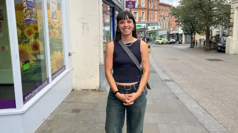 Woman with brown hair, purple top and jeans stands with her hands clasped in front of her and smiling on Stroud High Street.