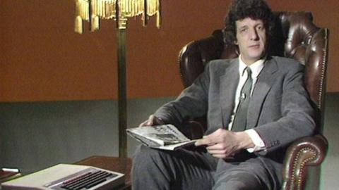 Presenter Chris Serle sitting in a high-backed chair with a newspaper on his knee and a keyboard on a table beside him.