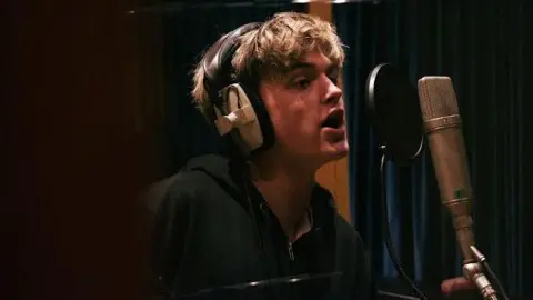 Oscar Hartland Oscar in a music studio singing in front of a microphone. He has headphones on and has short blonde hair and a black jacket on.