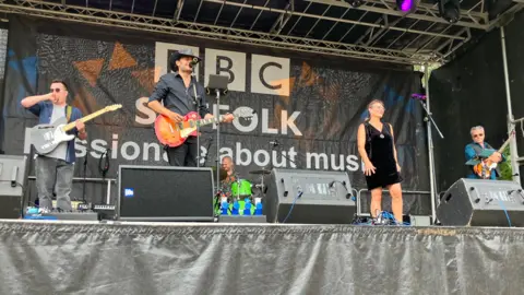 Ollie Ward/BBC A band performs on a small stage at a music festival. Three men play guitars while there is also a woman in front of a microphone. Another man is at the back playing the drums.