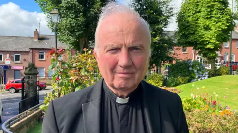 Bishop of Derry Donal McKeown has grey hair and is dressed in black. He is standing in front of flowers and trees in Brooke Park in Londonderry