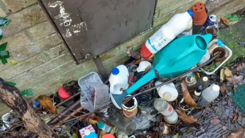 LDRS Rubbish at the door of a property. There is garden waste, plant pots, bottles, a watering can, bottles and cans.