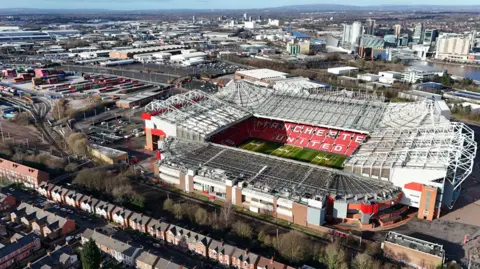 PA Media aerial drone shot of Manchester United's ground, with terrace houses in foreground, industrial Trafford Park to the left and skyscrapers and Pennine hills in background