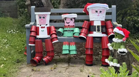 BBC/Amber Gash Three statues made of flowerpots sit on a bench. Two of them are depicting Father Christmas and Mrs Claus in red and white costumes. A smaller statue, dressed as an elf has been placed in between them. A fourth statue, a green coloured Grinch, is visible on the right of the image.