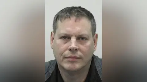 Mugshot of Steven Gauci. He is clean shaven with short dark hair tinged with grey and wearing a black and grey jacket.