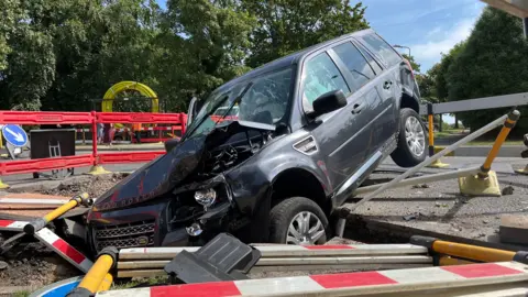 A Land Rover vehicle with its front stuck in a trench in the ground. Its bonnet and front lights have been damaged. Plastic fences that had been in place around the trench have been damaged and pushed over during the collision.