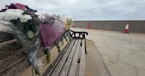 BBC/JIM SCOTT Bouquets of flowers attached to the back of a wooden bench, on a concrete esplanade by the sea.