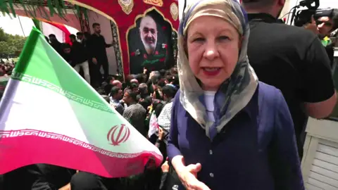 Lyse Doucet in a blue shirt and headscarf, next to a crowd of people and a large flag