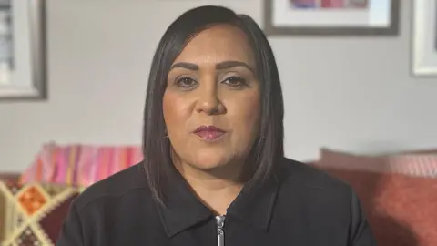 A head and shoulders photo of Ekta looking at directly at the camera. She is sitting on a sofa in a living room. She has straight dark shoulder-length hair and a black top with a collar and zip at the neck.