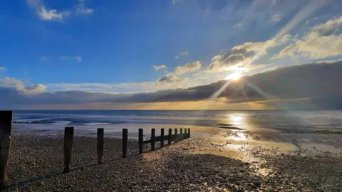 BBC Weather Watchers / Gadabout The pebbled beach has wooden posts in it that lead to the sea. There is a line a grey clouds in the sky and the sun is poking out above it creating rays of light. Above that the sky is blue.