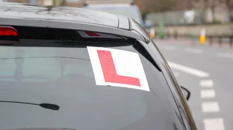 The back of a car on a road. It has a red L stuck to the window.