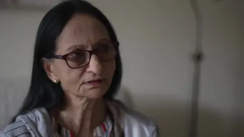 Yash Bandit, a woman with long dark hair wearing glasses, sitting in her apartment.