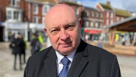 Alex Dunlop/BBC A man with a bald head, wearing a black suit jacket and blue tie, white shirt, stands in a market place.