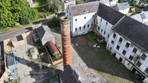 An aerial view of an old courtyard of a mill building with white industrial buildings gathered round an old brick chimney