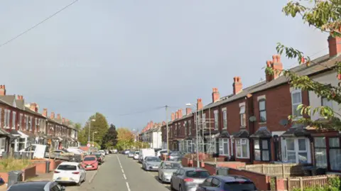 Residential street lined with red-brick terraced houses on both sides, with cars parked along the road. A clear roadway extends into the distance beneath an overcast sky, with trees, utility lines, and scaffolding visible among the houses.

