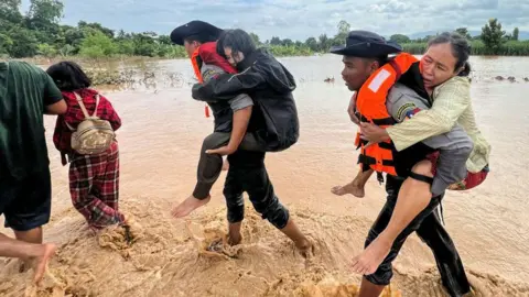 Getty Images Two male police officers carry women on their backs through muddy flood waters