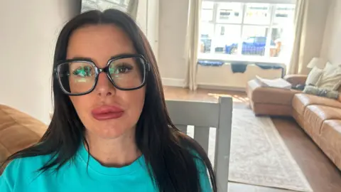 Nicola Austin-Ratcliffe looking into the camera which a straight face and pouting lips. She has long, straight dark hair and is wearing large, square glasses. She is sitting in a living room with a tan leather sofa and is wearing a turquoise t-shirt. 