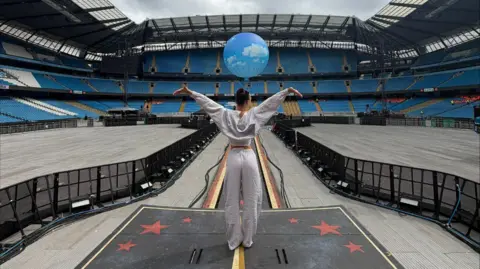 Ellis Grover Millie Harvey looks out to the empty stadium with her arms raised. She is wearing a white tracksuit and her hair is tied up in a bun.