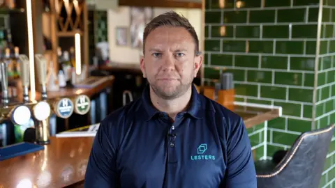 A man sat at the bar of a pub. He is wearing a blue polo shirt. The bar has green brick like tiles behind it. 
