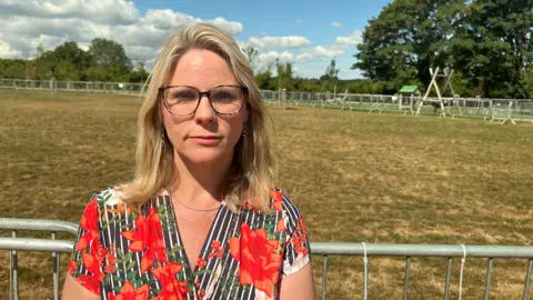 A picture of Michelle Hyde, a woman with shoulder-length blonde hair and glasses. She is wearing an orange, black and white floral top and she is standing in front of a metal fence on an area of fairly dry grass.