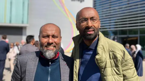 Jon Wright/BBC Mohammed Alam, in a blue fleece and grey suit jacket, and Mojlum Khan, wearing a khaki green coat, stand outside smiling at the camera.