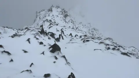 FELL TOP ASSESSORS A snowy summit of Helvellyn which shows black rocks peaking through. It is white-out conditions with poor visibility. 