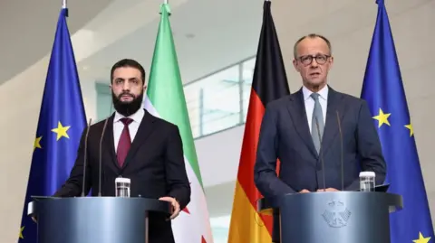 German Chancellor Friedrich Merz and Syrian President Ahmed al-Sharaa wearing suits and standing at podiums, in front of flags, hold a press conference at the Chancellery in Berlin, Germany