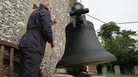 A man in overalls pulling a chain which is attached to a large church bell that is being hoisted above a church