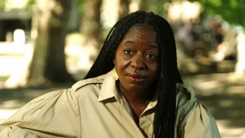 BBC A portrait photograph of a woman with black hair in braids, with blurred trees and grass in the background
