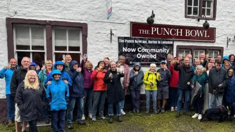 Management Team, Punch Bowl Inn, Burton in Lonsdale A mixed gender group of people in raincoats stand outside the Punch Bowl inn, raising glasses.