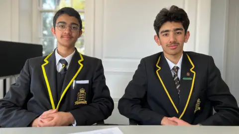 Yahiya and Ibrahim sit side by side at a table in a bright room with white walls and large windows. Both are wearing matching black school blazers with yellow trim, white shirts, and ties. Yahiya has a school crest and a white pocket square visible on the blazer. Their hands are resting on the table, and a sheet of paper lies in the foreground. 