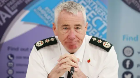 PA Media Jon Botucher sitting at a desk in front of a microphone with his hands clasped before him. He is wearing police uniform