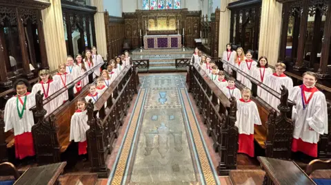 Ipswich Minster A group of young choristers within a church all wearing matching white and red robes. They are standing within the church's choir stalls toward the front of the building. 