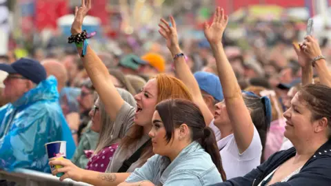 A crowd of people at Summer Fest at the Beach in Weston-super-Mare. Two girls can be seen in the centre of the shot. One has her arms up in the air, while the other is leaning against the barricade. Other punters can be seen around them.