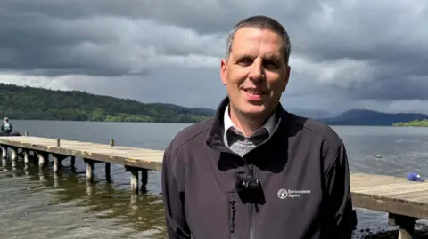 Andy Brown, water regulation manager at the Environment Agency. He has shaved grey hair and is standing in front of a small pier on Windermere. He is wearing a blue fleece with the Environment Agency logo, over a grey jumper and white shirt. He is smiling at the camera.