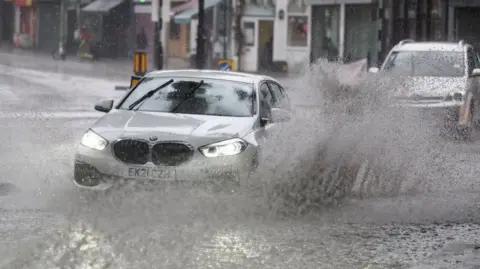 A silver car drives through water on the road in suburban London, creating a large splash. 