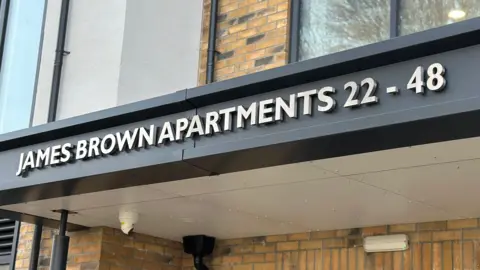 The front of a section of the new apartment block, which has light coloured brick work along with lettering that says James Brown Apartments 22-28 in silver on a black background.