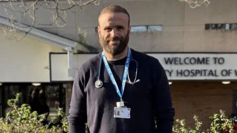 Jamie Roberts stood out the UHW Hospital in Cardiff. He is smiling wearing a navy blue jumper with his lanyard and stethoscope around his neck.