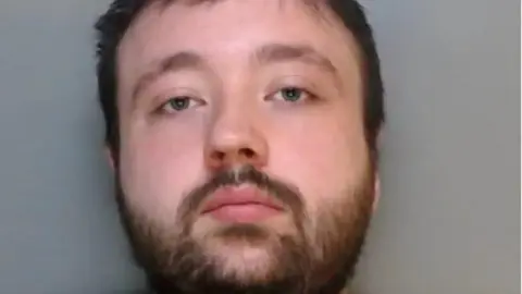 A mugshot of Cameron Greaves, who has a beard and moustache, short brown hair and blue eyes.