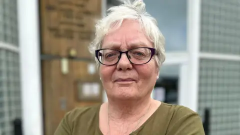 John Nurden A woman with black glasses and a cross necklace looks at the camera