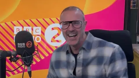Paddy McGuinness laughs in the Radio 2 studio. He wears glasses and a checked blue shirt