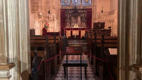 BBC/Richard Williams A small chapel seen from a carved stone doorway, with rows of wooden benches, stained glass window and an altar. It is roped off. Brown and white chequered tiles are on the floor.